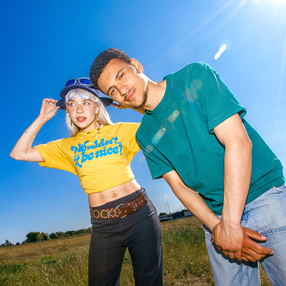 picture of artist Pixey in a yellow t-shirt standing next to a man in a green t-shirt and jeans standing in a field of grass on a sunny day