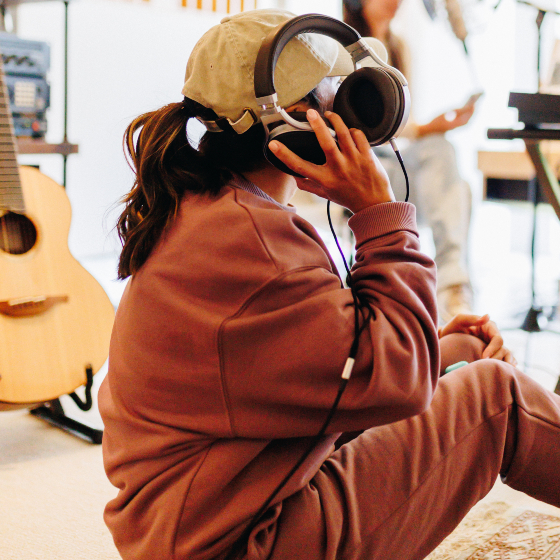 A woman sitting on the floor in a music studio holding headphones to one ear.