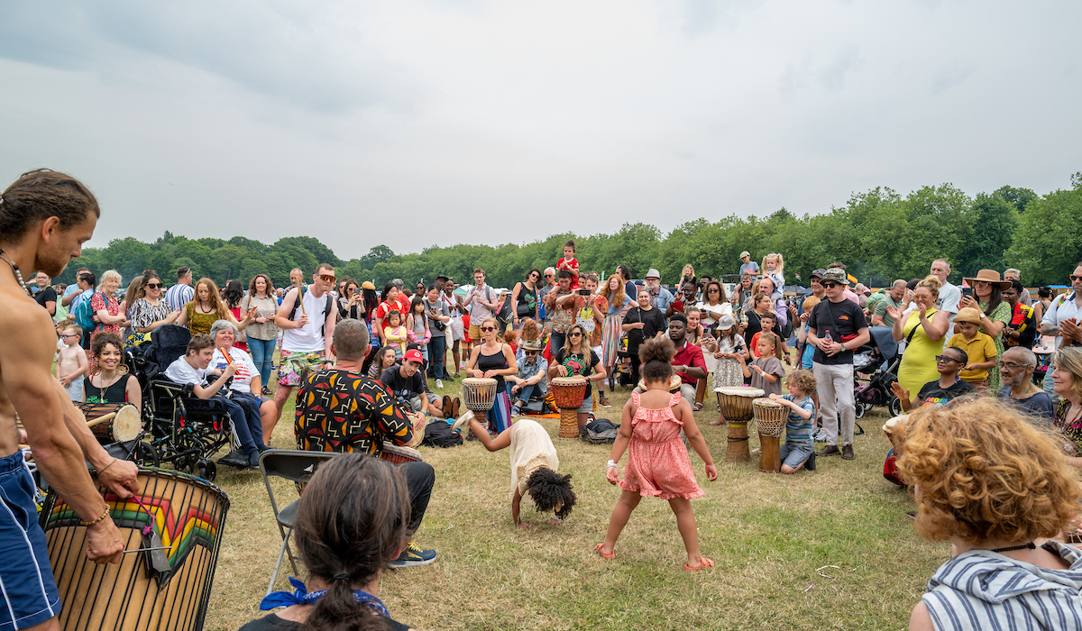 A group of people gather in Sefton Park at Africa Oye Festival, engaging in a lively drum circle and dance performance, surrounded by an enthusiastic audience on a sunny day.