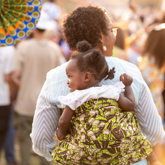 A person carries a toddler on their back at a bustling outdoor festival