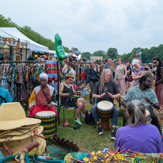 At Africa Oye festival, people sit, playing drums in the foreground. Stalls with colorful clothing and hats in the background amidst a grassy park setting.