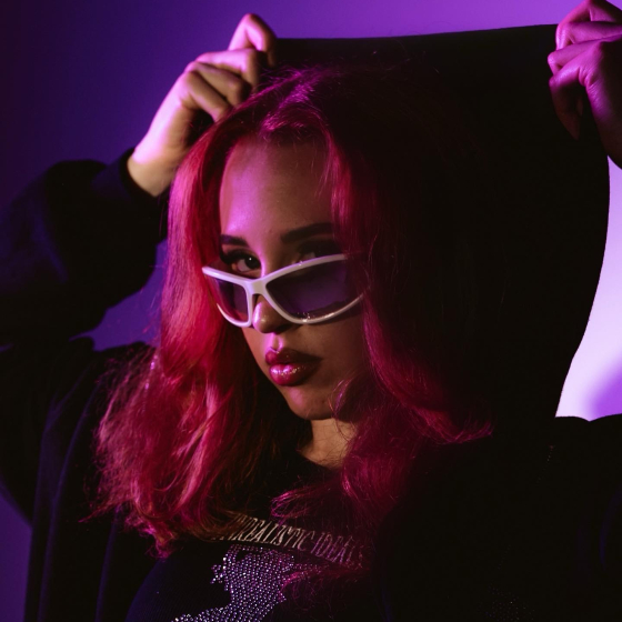 Person wearing a hooded top and white sunglasses, standing against a purple-lit background. 