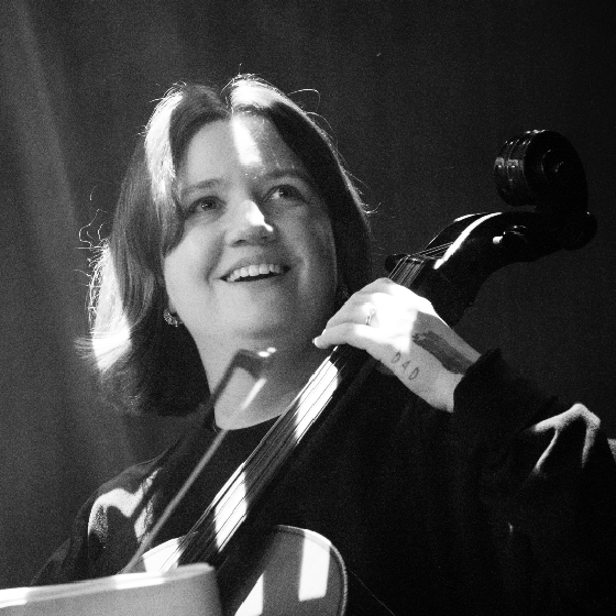 A musician smiling while playing a cello on stage, under soft stage lighting.