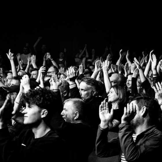 Black and white photo of a crowd at a concert, with several individuals raising their hands enthusiastically. There's a sense of excitement and movement in the scene.