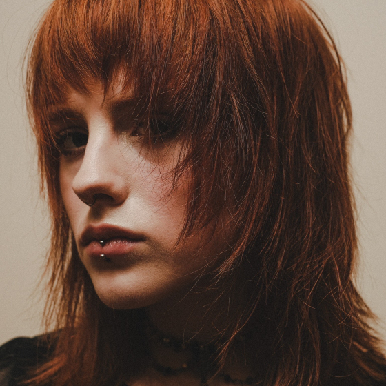 Close-up portrait of a person with red hair and a lip piercing, set against a neutral background.