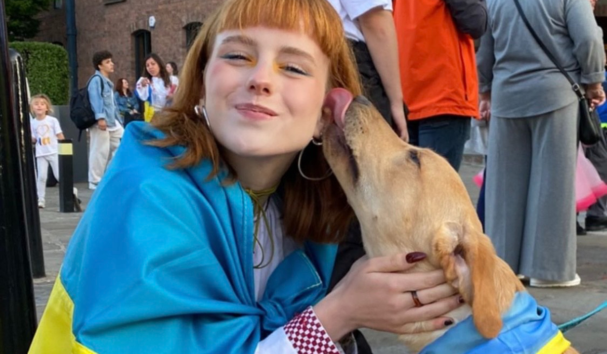 Person with a Ukrainian flag over their shoulders sitting and smiling, being affectionately licked on the face by a dog.