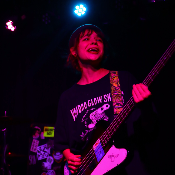 Musician performing on stage with a bass guitar, wearing a Voodoo Glow Skulls T-shirt, under colorful stage lighting.