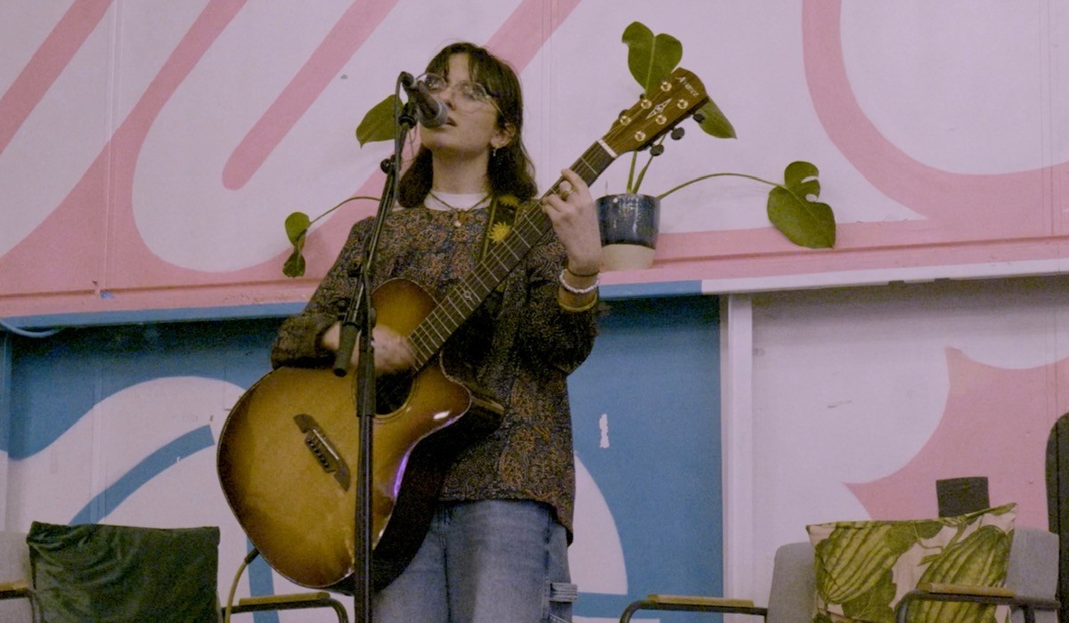 A musician performs on stage, holding an acoustic guitar, in front of a pastel-colored mural featuring abstract swirls. The performer wears glasses and a patterned shirt, and is singing into a microphone.