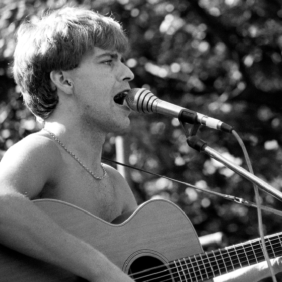 Black and white photo of a musician from The La's playing an acoustic guitar and singing into a microphone outdoors.