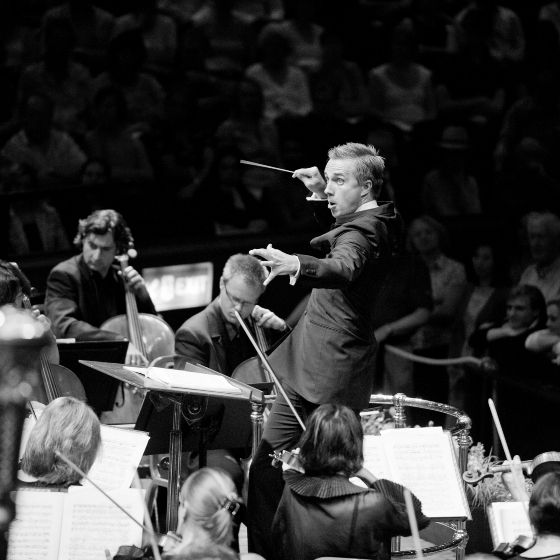 A person conducting the Royal Liverpool Philharmonic orchestra at a BBC Proms concert, focusing intently while musicians with various instruments perform around them.