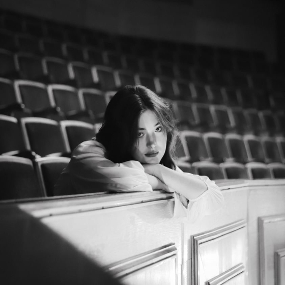 Person resting their chin on their hands, leaning over the back of a theater seat, looking thoughtfully towards the camera. The setting is a dimly lit auditorium with rows of empty seats. The photo is in black and white, emphasizing a moody and contemplative atmosphere.