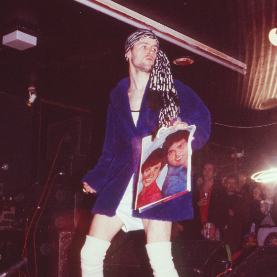 An 80s photo of a man in a blue coat and head scarf bandana, holding a poster exuding confidence whilst in a night club.