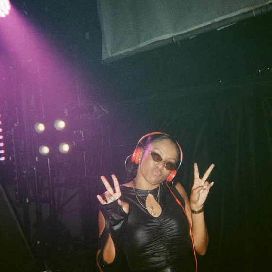 Person with headphones and sunglasses making a peace sign gesture, standing under stage lights in a dimly lit setting.