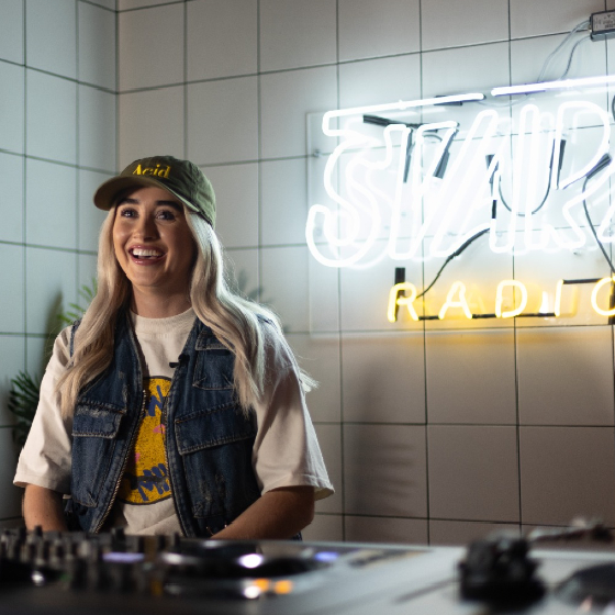 A woman smiling in front of a white tiled wall with a neon sign.