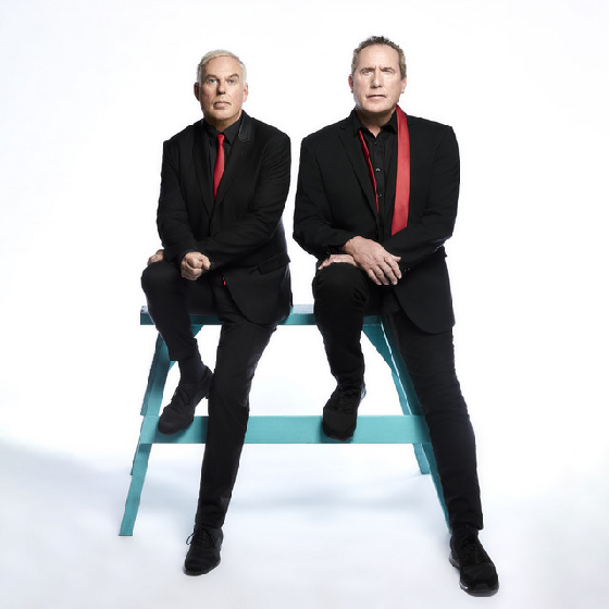 Two men both wearing black suits and a red tie, stood on a blue bench against a whte background