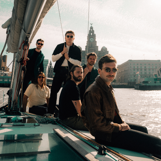 A group of six men sit on a boat with the iconic Liverpool Liver Building in the background on sunny day