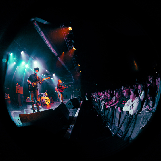 A fish-eye photo of a band playing on stage with people from the crowd against the barrier