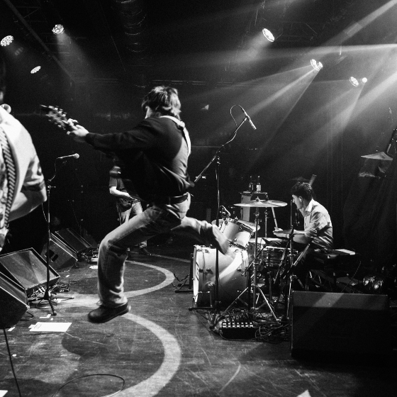 A black and white image of a band playing on stage, the guitarist jumping in the air