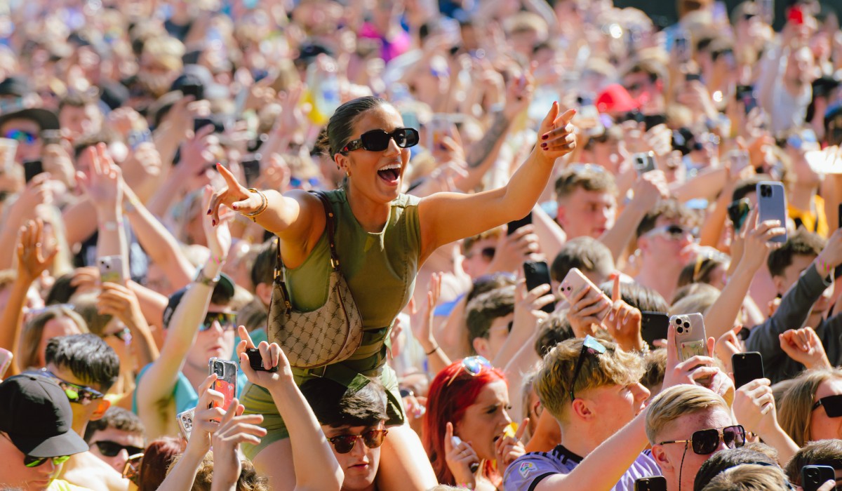 A girl on someone's shoulders in a crowd at a lively outdoor music event on a sunny day