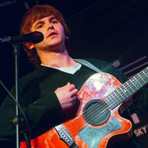 A person standing on a stage playing an acoustic guitar with a red and orange marbled finish. The person is positioned behind a microphone stand and is wearing a dark long‑sleeved top layered over a lighter shirt. The background is dimly lit, with indistinct equipment and stage elements visible.