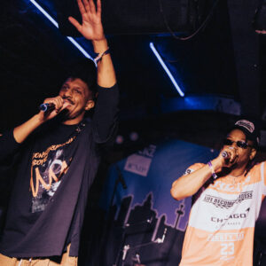 Two performers are on stage in a dimly lit venue, each holding a microphone. One performer on the left is wearing a long‑sleeved black shirt with a graphic design and has one arm raised. The performer on the right is wearing a light-colored T‑shirt with printed text and a cap, holding the microphone close to their mouth. Blue and purple stage lighting highlights the scene, and a backdrop featuring a city skyline silhouette is visible behind them.