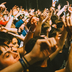 A densely packed crowd at a live music event, with many raised arms and hands pointing upward. People are standing close together, wearing festival-style wristbands and casual clothing. The lighting is warm and dim, suggesting an indoor concert atmosphere. The scene captures the high energy and movement of the audience as they engage with the performance.