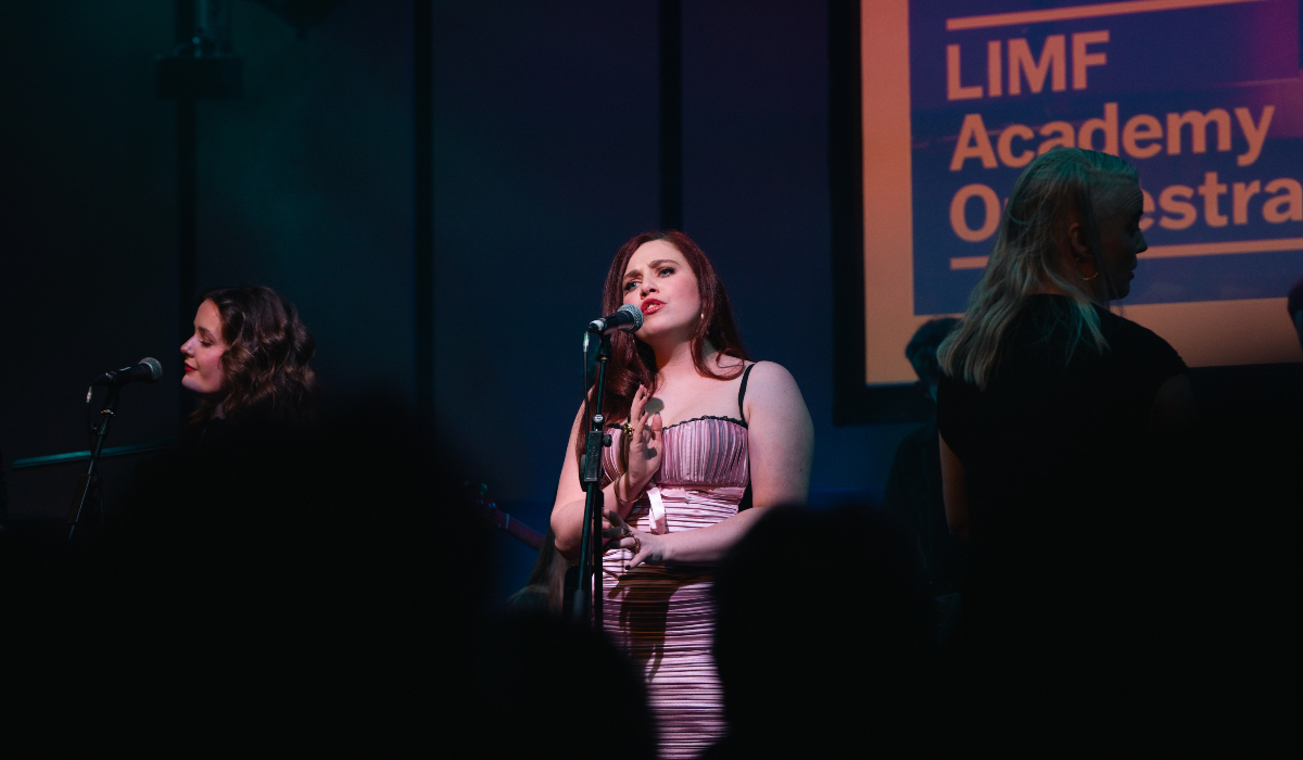 Faye Donna Francis stands at a microphone on a dimly lit stage, wearing a pink satin dress, with other performers partially visible to either side. A screen in the background displays the text 'LIMF Academy Orchestrated' and audience silhouettes are visible in the foreground.