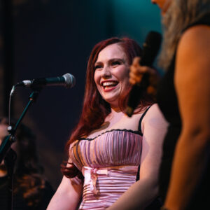 Faye Donna Francis standing on a stage behind a microphone, wearing a pink strapless dress with a bow at the waist. Another person holding a microphone is partially visible to the right, with stage lighting and a dark background.