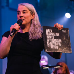 Katie Chatburn on a stage holding a microphone and a black square book reading ‘WHEN INSPIRATION DRIVES INTERVENTION RESULTING IN ELEVATION - LIMF Academy’. Blue stage lighting and an orchestra are visible in the background.