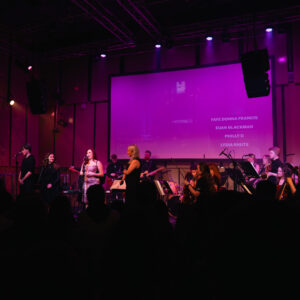 A wide shot of a live orchestral performance on stage under purple lighting, with multiple singers at the front and a seated orchestra behind them; a large projection screen above the stage displays performer names while audience silhouettes appear in the foreground.