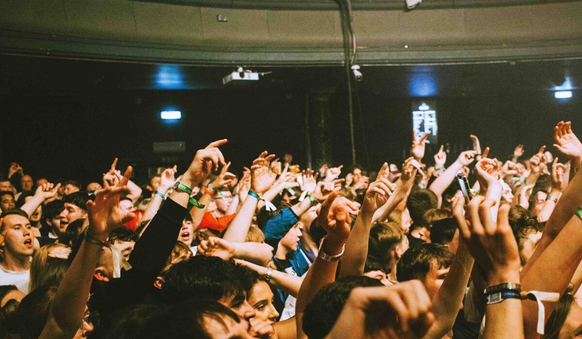 A dense crowd at an indoor live music venue, with many audience members standing close together and raising their hands, some wearing wristbands, while a balcony with more people is visible above the main floor under warm, dim concert lighting.