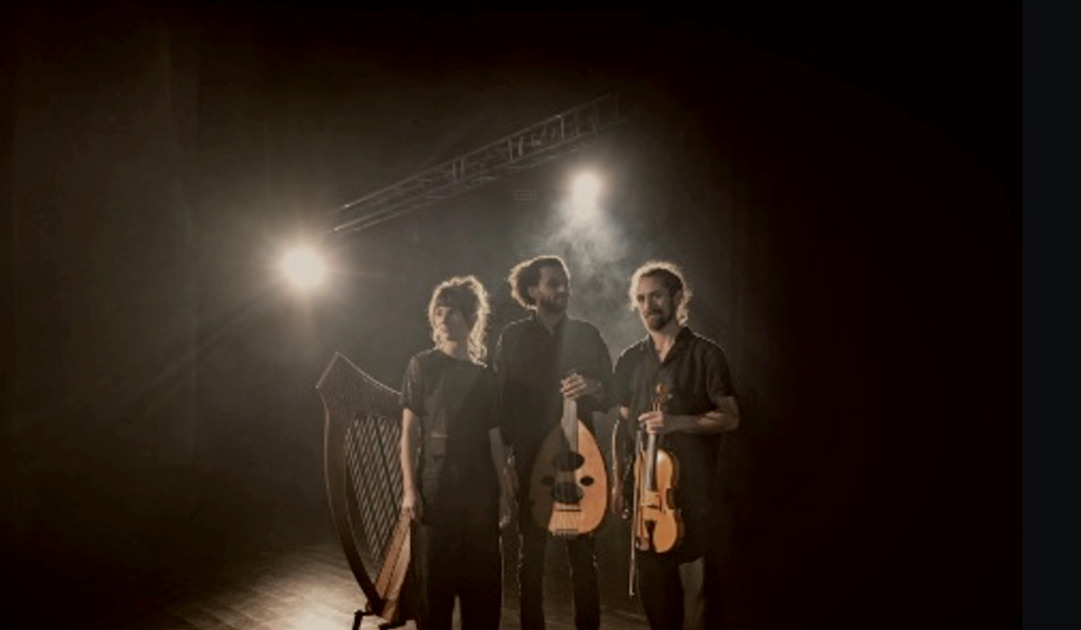 Three musicians stand on a dimly lit stage, facing forward under bright backlighting and light haze. Each person is dressed in dark clothing and holds or stands beside a traditional string instrument: a large harp positioned to the left, a lute held in the center and a violin held on the right. Strong stage lights behind the group create silhouettes and soft highlights around the figures, with metal rigging faintly visible overhead. The background is mostly dark, emphasizing the performers and their instruments.