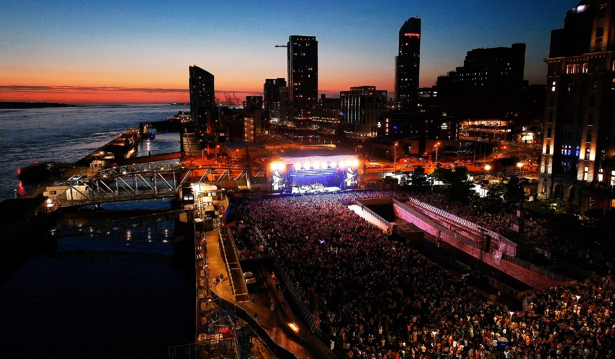 Liverpool waterfront skyline at dusk with a large outdoor concert crowd gathered in front of a brightly lit stage beside the water.