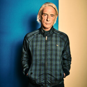Person in a green plaid zip-up jacket standing against a blue and beige split background, facing the camera with hands in pockets.