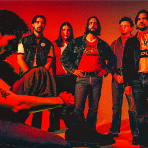 Six people in a studio pose against a vivid red-orange background; five stand in a line wearing denim and casual clothing, while one sits in the foreground adjusting a black lace-up boot. Dramatic red lighting creates a gritty, rock-style look, and a central T-shirt shows partially visible text including 'Party' and 'F***ing America'.