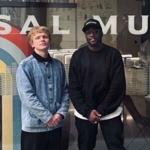 Two people stand outside a glass-fronted storefront with large white lettering, wearing casual dark and denim outfits with hands clasped in front of them.