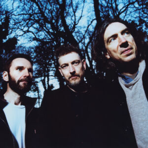 Three people standing outdoors in front of leafless trees, looking in different directions under cool blue-toned lighting.
