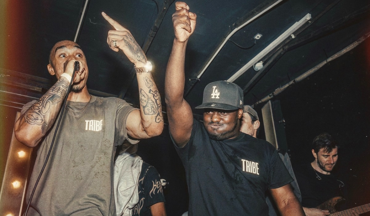 Two performers in TRIBE T-shirts gesture to the crowd during a live music performance, with musicians and stage lighting visible in the background.