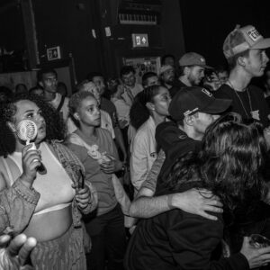 Black-and-white photo of a crowd packed into a live music venue, watching a performance. One attendee in the foreground holds a small handheld fan while others stand closely together near the stage.