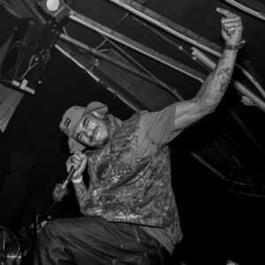Black-and-white photo of a performer holding a microphone and raising one arm during a live music performance in a small venue.