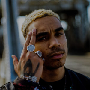 Close-up portrait of a person with short bleached hair, wearing silver rings and a bracelet, with a hand raised to the forehead against a blurred outdoor background.