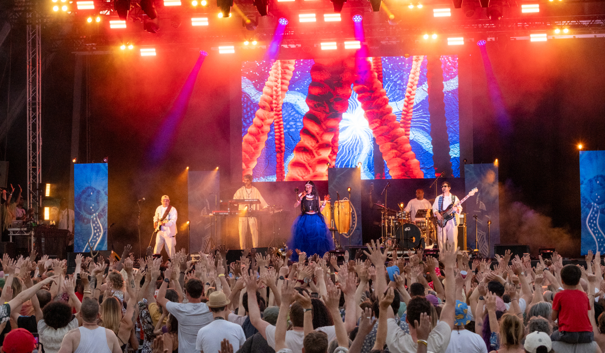 Large crowd with raised hands watching a live band perform on a brightly lit festival stage with colourful visuals on a giant screen.