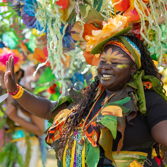 A person in a vibrant festival costume raises one hand while standing amid colourful decorations. The outfit features layered green leaf-like materials, patterned fabric in bright orange, yellow and green tones, face paint with gold accents, and a large floral headdress made from oversized flowers and cascading ribbons. The background is filled with blurred festival participants and vivid multicoloured decorations in bright daylight.