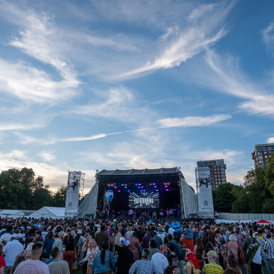 Large outdoor concert crowd gathered in front of a festival stage at dusk, with colourful stage lighting and a large video screen displaying a performer. White festival tents and trees surround the venue, while dramatic wispy clouds stretch across a bright blue evening sky above. Multi-storey buildings are visible in the background.