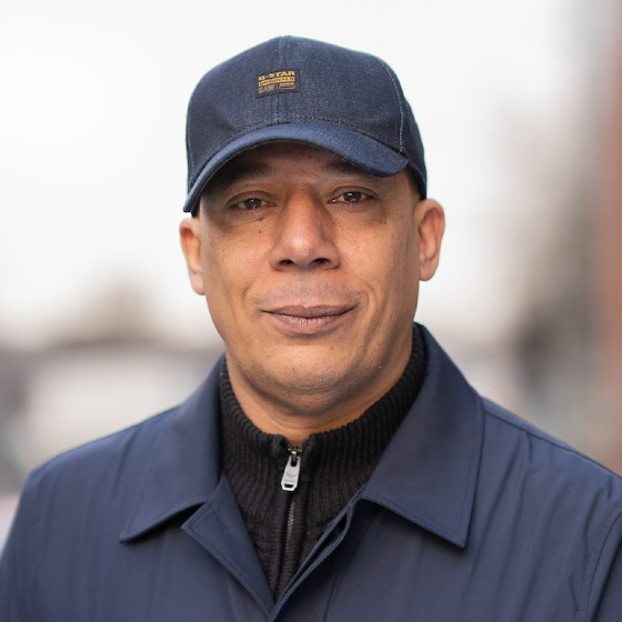Head-and-shoulders portrait of a person wearing a navy blue baseball cap with a small logo patch, a dark zip-up knit top, and a blue jacket. The person is facing the camera against a softly blurred outdoor background with neutral tones.