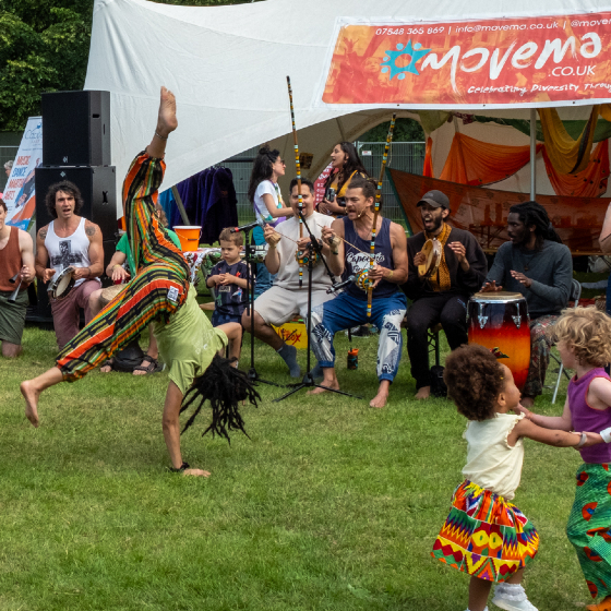A lively outdoor festival performance on a grassy field. In the foreground, a performer in a brightly striped outfit balances in a one-handed handstand while musicians seated behind play percussion instruments and shakers under a white canopy. Several children dance and play nearby. A banner above the stage reads 'Movema.co.uk', and trees surround the event area in the background.