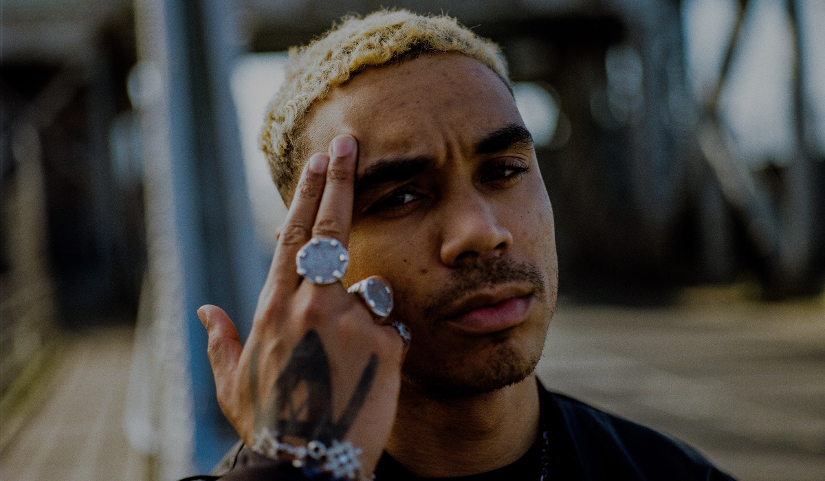 Close-up portrait of Koj with short light-coloured hair, resting two fingers against the side of his forehead. He is wearing several silver rings and a bracelet, with a dark tattoo visible on the back of his hand. The background shows an outdoor wooden bridge with structural beams blurred out of focus.