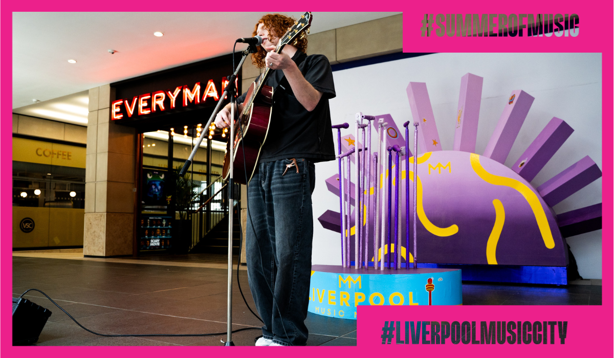 A person with a guitar singing at a microphone with a pruple art installation behind them.