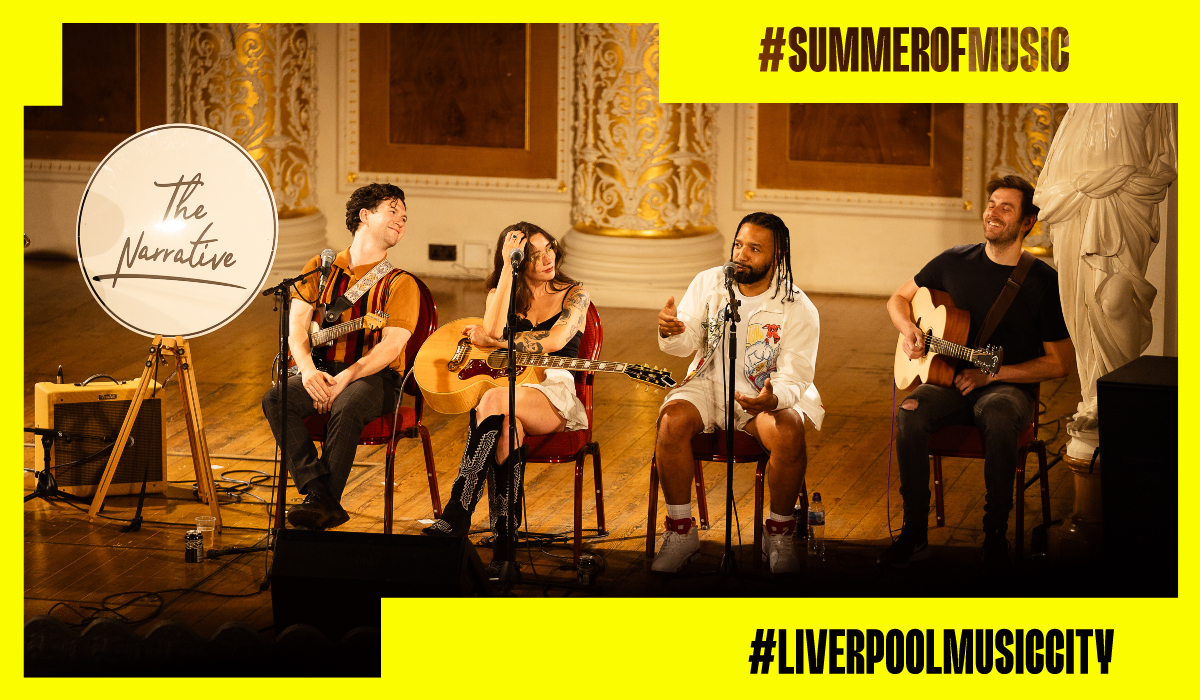 Four musicians sit on a stage during an acoustic performance in an ornate hall with gold decorative columns and wooden flooring. Two performers hold guitars while a third speaks into a microphone. A round sign on an easel reads 'The Narrative'. The image is framed with a bright yellow graphic border featuring the hashtags '#SUMMEROFMUSIC' and '#LIVERPOOLMUSICCITY'.