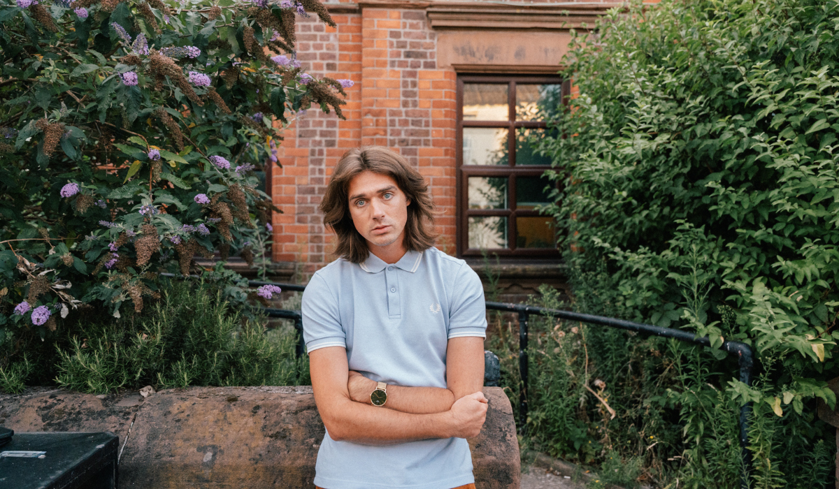 A person wearinf a light grey t-shirt stood with their arms folded in front of a brick building.
